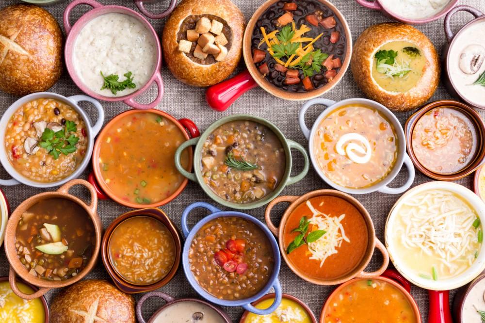 High Angle View of Various Comforting and Savory Gourmet Soups Served in Bread Bowls and Handled Dishes and Topped with Variety of Garnishes on Table Surface with Gray Tablecloth